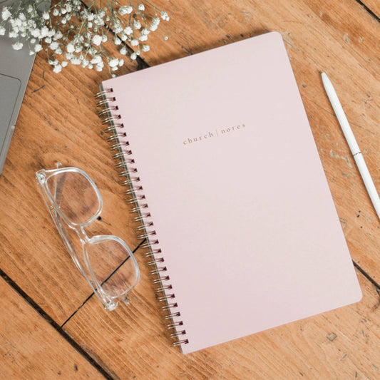 Pink notebook with 'church notes' on a wooden surface with glasses and a pen.