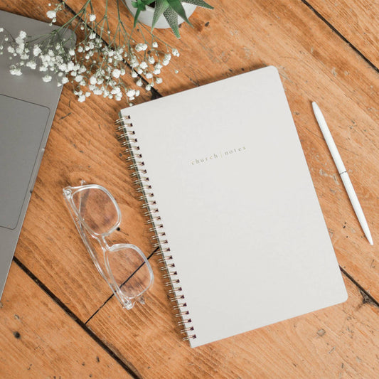 Notebook, glasses, pen, and laptop on a wooden surface with flowers.