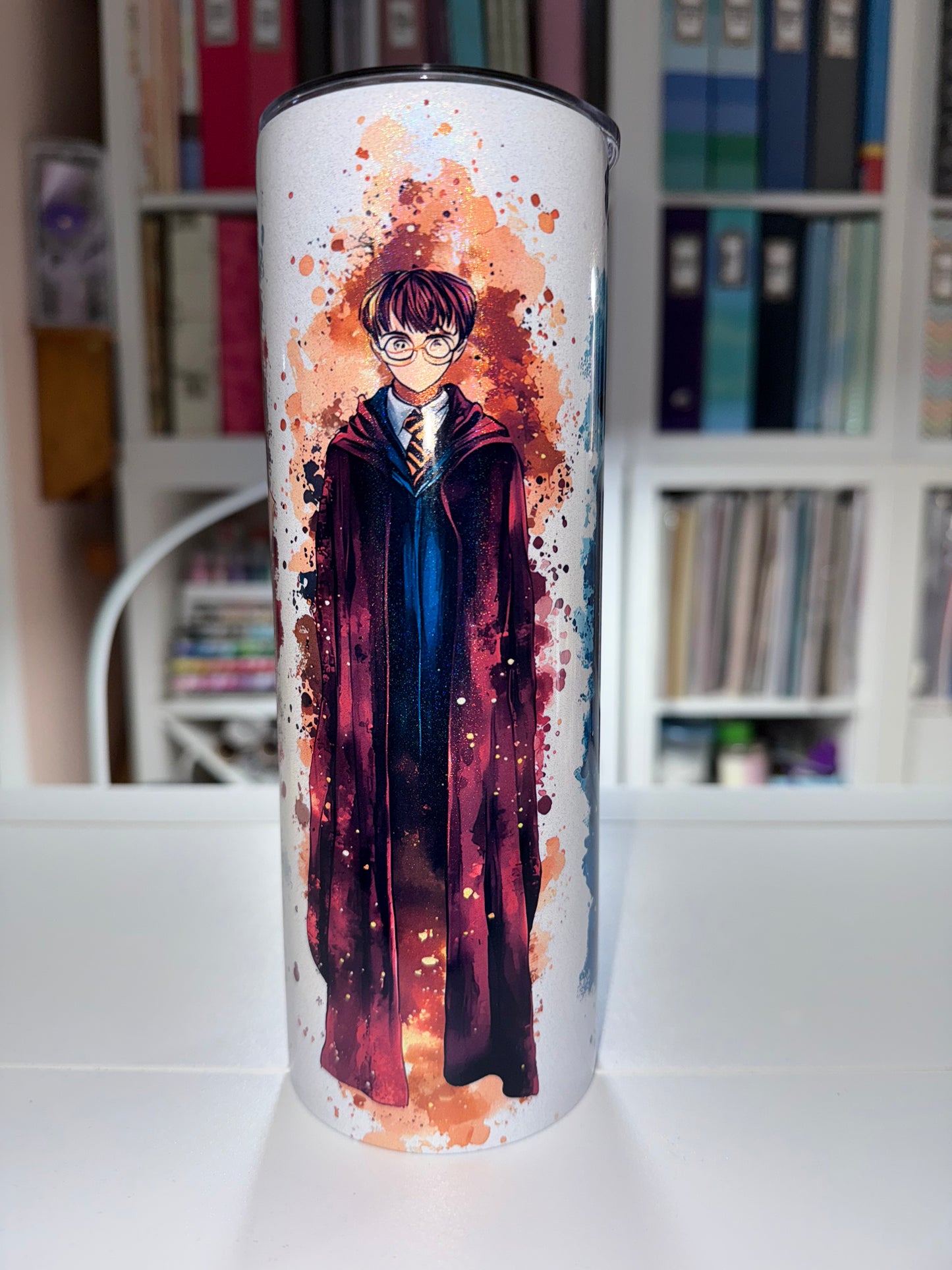 Tumbler with artistic design of a person lying down on a colorful background, placed on a surface with a blurred bookshelf in the background.