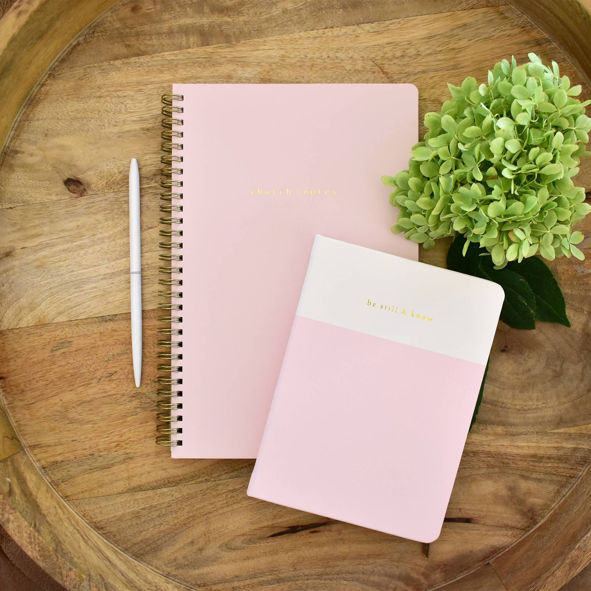 Two pink notebooks on a wooden surface with a pen and green flowers.