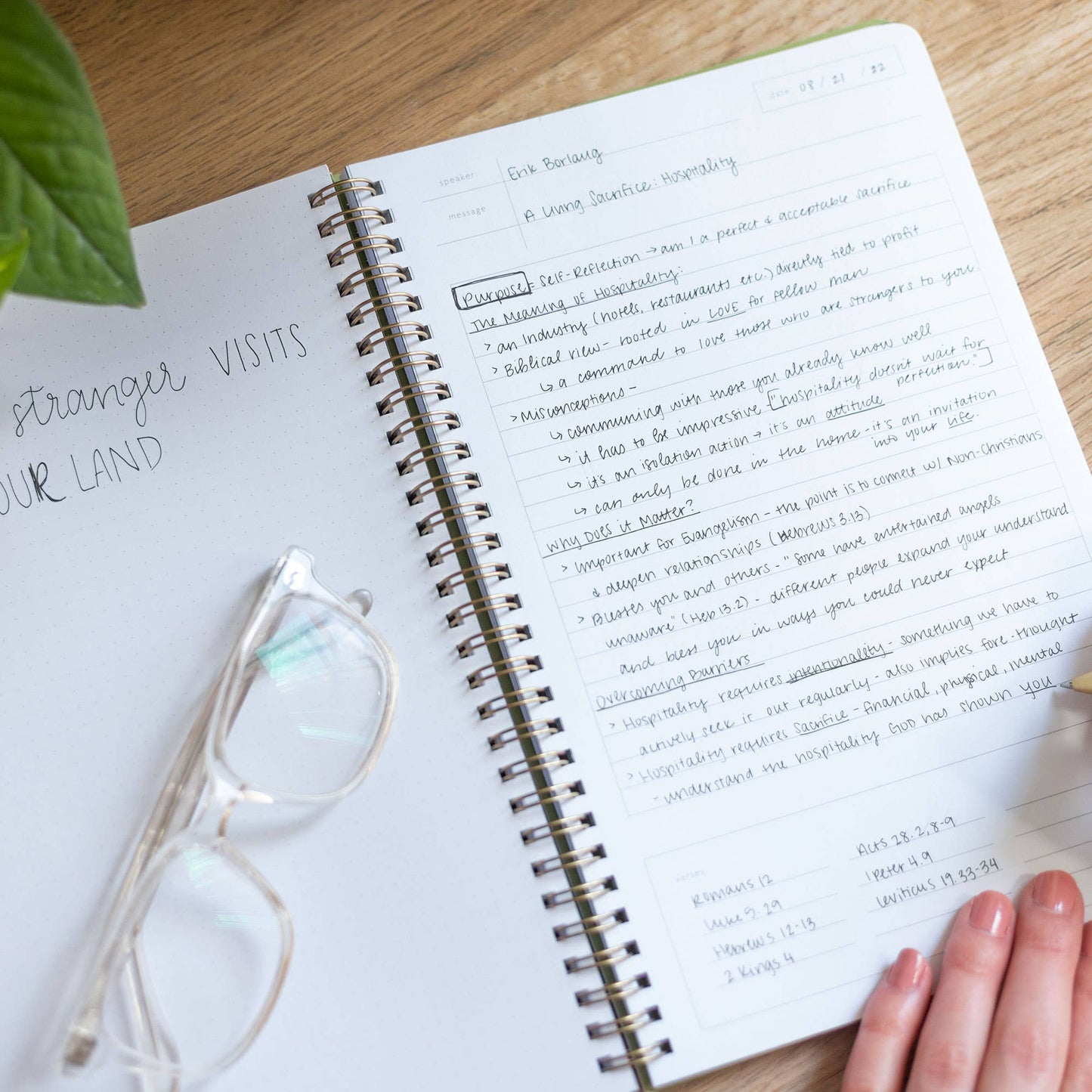 Open notebook with handwritten notes on a wooden surface, with glasses and a plant nearby.
