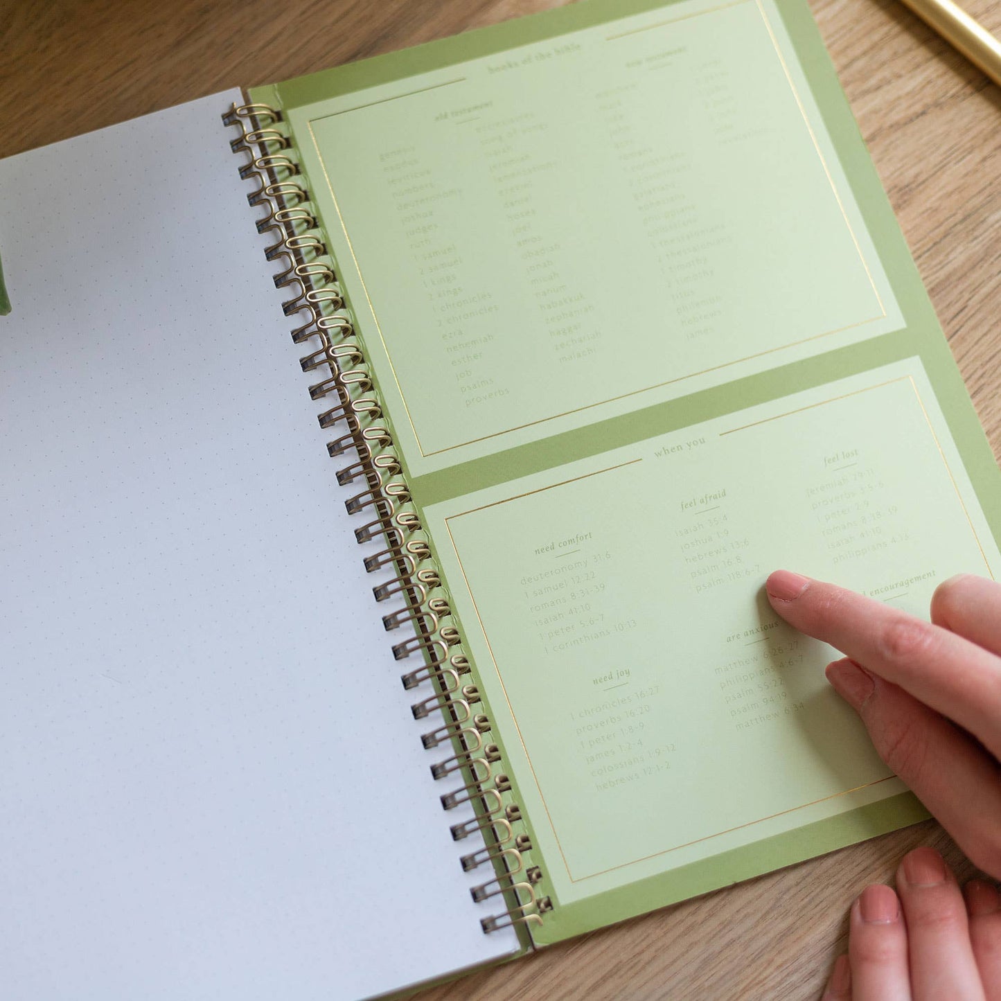Spiral-bound planner with green cover on a wooden surface, hand pointing to text.